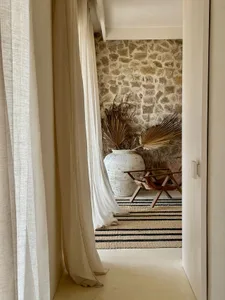 A neutral-toned living room features a stone wall, a large vase with dried palm leaves, a wooden chair, and a striped rug und