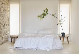 A minimalist bedroom featuring a bed with white cotton bedding, two windows with sheer curtains, and rustic wooden stools wit