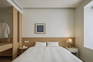 A minimalist white bedroom with a wooden headboard, white bedding, and a nightstand with a lamp.