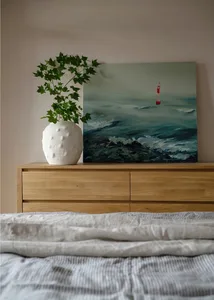 A bedroom with a wooden dresser, a green plant in a white vase, and a painting of a person in the ocean.