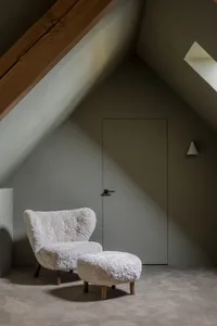 A cozy mid-century living room with a white wool armchair and matching ottoman, set against a muted green wall and slanted ce