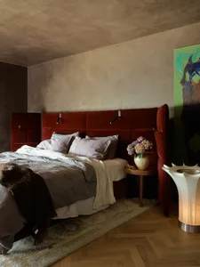 A minimalist bedroom featuring a red velvet headboard with integrated lighting, a wooden nightstand with a vase of flowers, a