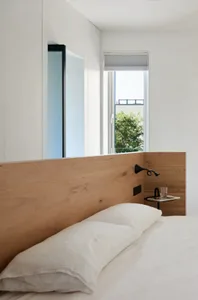 A minimalist bedroom with a wooden headboard and white bedding, featuring a side table with a glass and a newspaper.