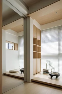 A minimalist living space featuring wooden elements and a sunlit room with a bookshelf and window.