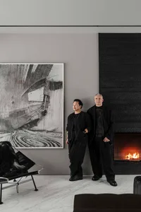 Two individuals standing in a modern art museum with a large black and white abstract painting and a fireplace.