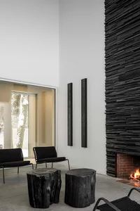 A modern black living room with exposed brick fireplace, birds-eye-view tree trunk stools, black woven rope chair, and large
