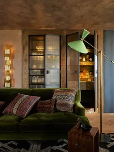 A modern living room with a green velvet sofa, patterned pillows, and a wooden floor, featuring a sleek floor lamp and a glas