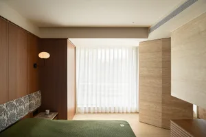 A modern minimalist bedroom with brown wooden finishes and white sheer curtains by the window.