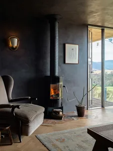 A cozy industrial-style living room features a concrete fireplace, wood floors, and a rustic rug, with a view overlooking a g