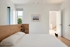 Modern minimalist bedroom with wooden headboard, white bedding, cityscape artwork, and black cat.
