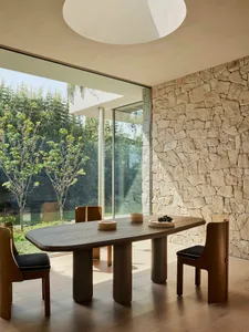 A minimalist dining room with a wooden table and a stone wall, featuring floor-to-ceiling windows that allow natural light to