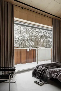 A minimalist bedroom featuring concrete flooring, wooden furniture, and a window with a view of snowy trees.