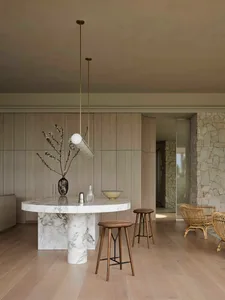 Modern minimalist kitchen with marble countertop and stools, featuring a tall cylindrical lamp and wooden chairs.