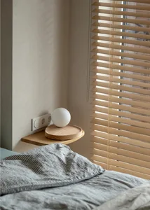 A minimalist bedroom with a wooden blind, grey sheets, and a round wooden tray with a white globe lamp.