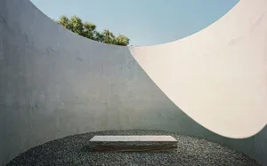 A minimalist concrete sculpture garden featuring a smooth, rectangular stone pedestal surrounded by pebbles and set against c