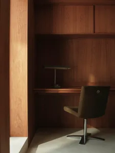 A modern home office with wooden furnishings, including a desk, chair, and lamp. The space is illuminated by natural light.
