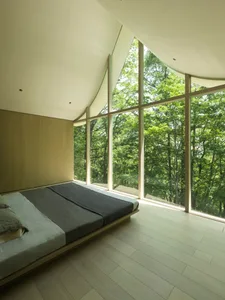 A modern minimalist bedroom with a large window showcasing a forest view, featuring wooden flooring and a low-profile bed.