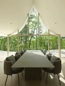 A modern, minimalist dining room featuring a long table surrounded by cushioned chairs, with large glass windows overlooking