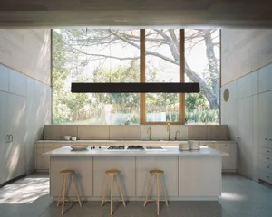 A modern minimalist kitchen featuring a large window, wooden stools at a central island, and a glass backsplash.