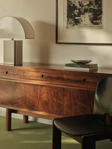 A modern office desk made of wood with a lamp, chair, and framed artwork.