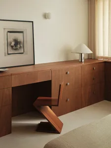 A modern minimalist office space featuring a wooden desk and chair with a wall-mounted light fixture and abstract artwork.