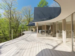 Modern residential deck featuring a dining table and chairs with wood flooring and glass railings surrounded by greenery.