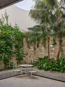 A modern outdoor space featuring a circular white table and chairs on a concrete patio surrounded by greenery and palm trees.