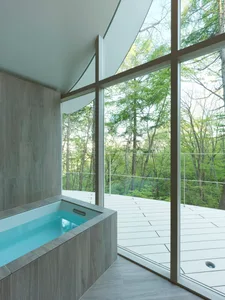 A modern residential bathroom with a glass wall overlooking a forest, featuring minimalist design and a wooden bathtub.