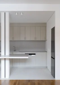 A modern, minimalist residential kitchen with light grey cabinets, a white countertop, and a backsplash made of small, square