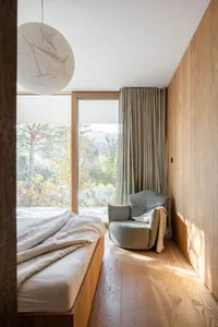 Modern minimalist bedroom with a wooden bed, a green armchair, and a large window overlooking nature.