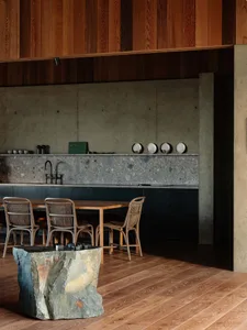A modern industrial-style residential kitchen featuring concrete countertops, stone accents, wooden dining table, metal chair