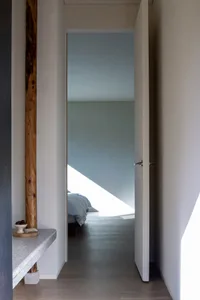 A minimalist bedroom with a white door, wooden floor, and minimal furnishings.