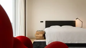 Modern minimalist bedroom featuring minimalist white fabric furnishings on a wooden bed platform, with a red curved chair and