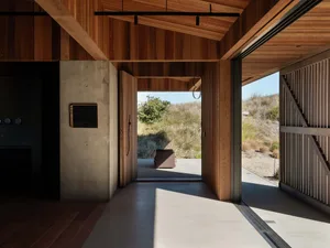 An open-plan minimalist entryway with wooden walls, floor, and ceiling, featuring a concrete partition and a view of the natu