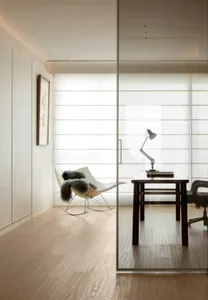 A modern home office featuring a glass wall, wooden desk, and comfortable rocking chair.