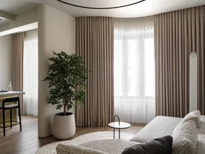 A modern living room featuring beige curtains, patterned fabric sofa, wood flooring, and a potted plant.