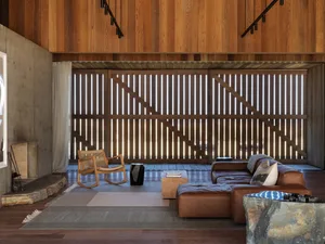 A modern living room with hardwood flooring, a leather sectional sofa, a wooden rocking chair, and a large window with vertic