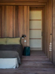 A modern minimalist bedroom featuring wood finishes, a bed with green pillows, and a small green stool.