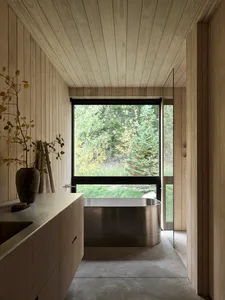 A minimalist bathroom featuring wood-paneled walls and a modern metal bathtub with a large window overlooking a forest.