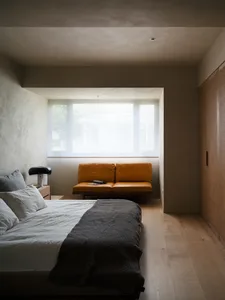 A minimalist bedroom featuring a bed with grey and white bedding, an orange lounge sofa, and wooden flooring, with a window l