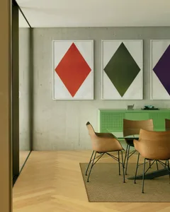 A modern dining room featuring wooden chairs, a glass table, metal legs, and geometric artwork on a concrete wall.