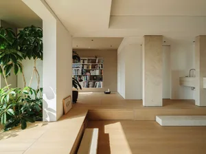 A modern living room featuring wooden flooring and concrete structural columns, with a bookshelf filled with books and potted