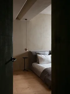 A modern minimalist bedroom featuring a bed with a low-profile fabric headboard, white bedding, and wood flooring.