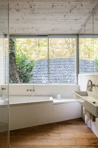 A minimalist bathroom interior with a white stone bathtub and sink, large window overlooking a forested area, and wooden floo