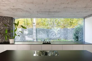 Modern kitchen interior with minimalist design featuring concrete countertops, large window, and potted plant.