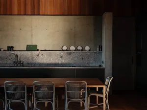 Modern kitchen with concrete countertops, wooden chairs, and dark wood paneling.