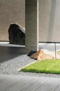 Modern landscape design featuring concrete patio, gravel pathway, and lush green grass with large rocks and a column.