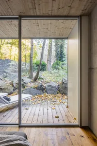 Modern minimalist doorway leading to a serene wooded area with leaves on the ground and a stone wall.