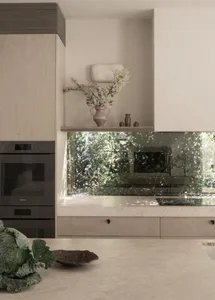 A modern, minimalist kitchen with taupe cabinets and a reflective backsplash showcasing greenery.
