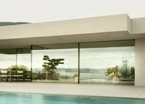 A modern residential exterior featuring a large glass wall overlooking a serene water body and distant hills.